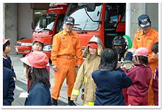 小学生が消防署を見学し、署員に話を聞いたり、消防服を着せてもらっている様子の写真