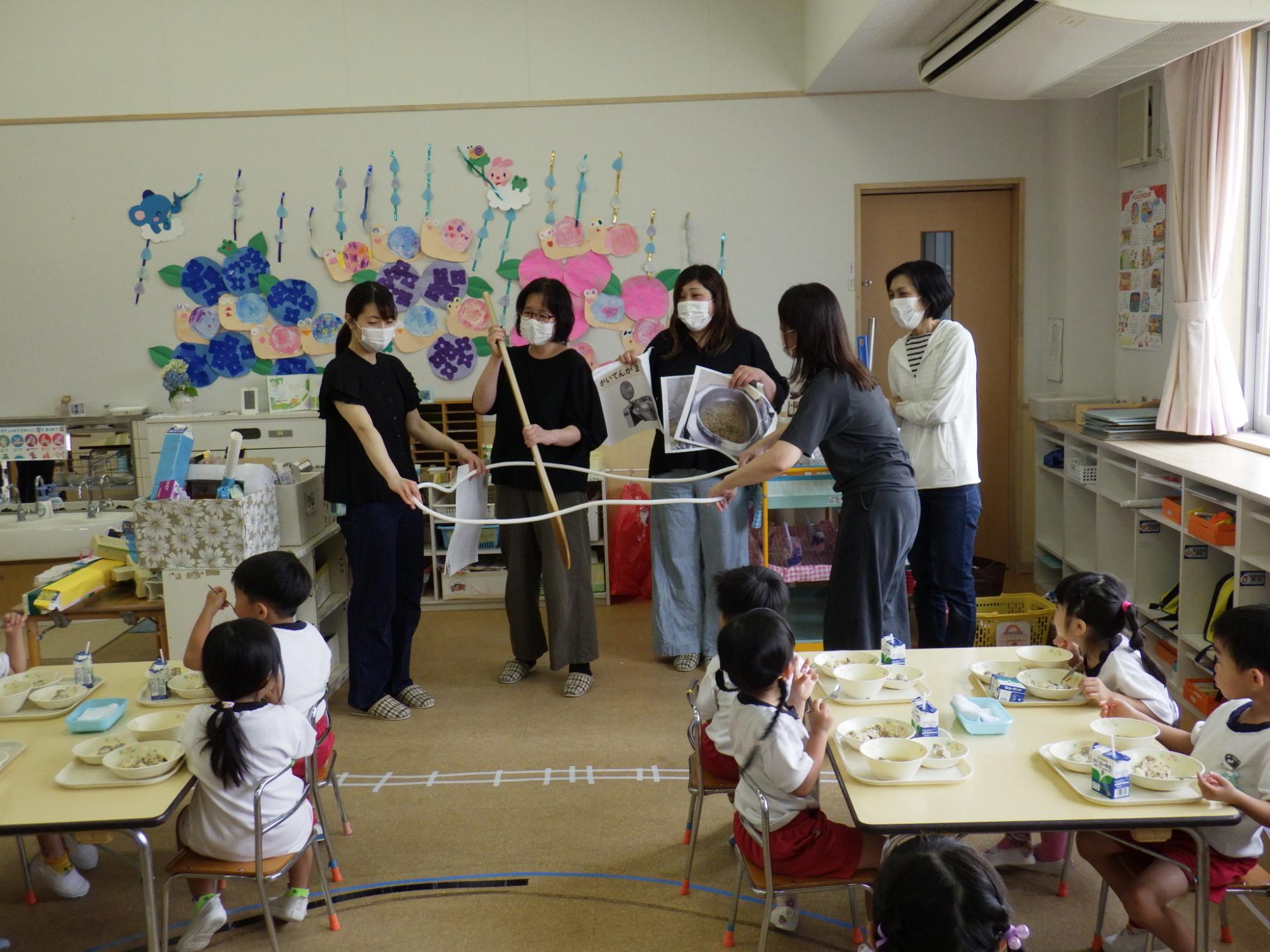 園児の前で、調理員とえいようしで、給食を作っている釜の大きさをロープで作って、大きなじゃもじで混ぜています