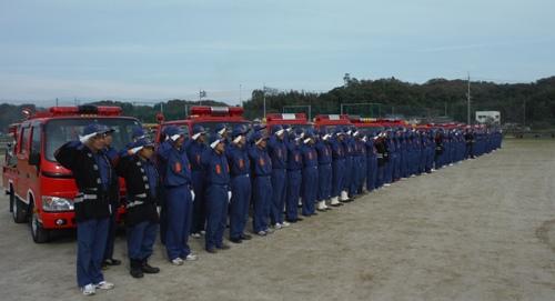 ずらりと並んだ消防車の前で整列する消防団員の写真