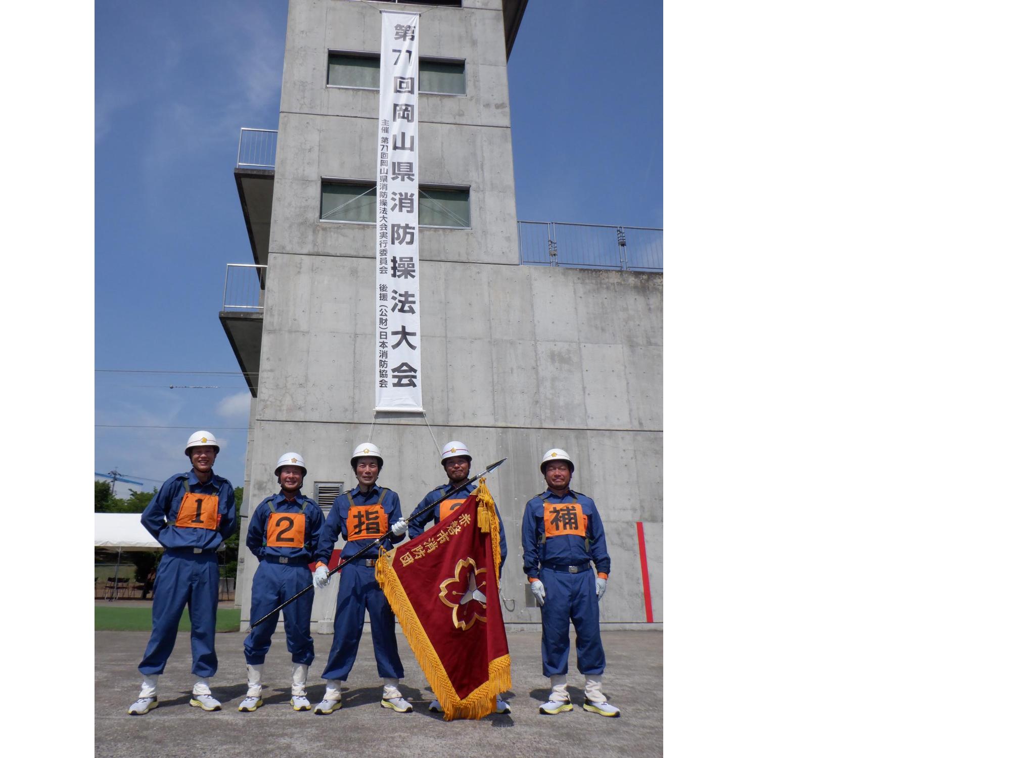 消防学校の訓練棟前で消防団旗をもった赤磐市消防団の代表選手が記念撮影